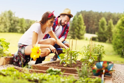 A young woman and man are working together in a well-maintained garden, planting or tending to a bed of young vegetable or flower plants. The woman, wearing a white t-shirt, plaid shorts, and a pink headband with a floral pattern, is leaning forward with a focused expression as she handles the plants. The man, dressed in a red and blue checkered shirt, white t-shirt, and a straw hat, is also engaged and smiling gently while supporting the gardening task. The garden bed is edged with wooden planks, and various garden tools and decorative plant markers are visible nearby. The soil appears rich and dark, and the young plants are emerging with bright green leaves, contrasting with the natural tones of the soil and wood. In the background, the garden opens to a lush, softly blurred landscape of trees and open fields, suggesting a countryside or suburban setting in Kensington or the surrounding area. The lighting indicates a clear, sunny day, enhancing the vibrant colours and natural textures, showcasing an inviting outdoor space suitable for gardening services provided by Lawn Mowing Kensington.