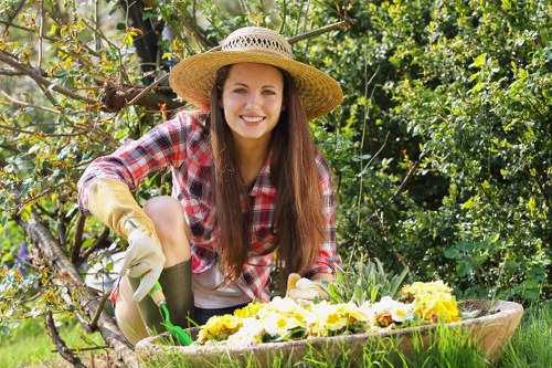 A young woman with long brown hair, wearing a wide-brimmed straw hat, a red and white checkered shirt, and gardening gloves, is kneeling in a lush garden during daylight hours. She is smiling at the camera while tending to a flowerbed filled with yellow and white blooms, possibly pansies or similar seasonal flowers. Behind her, the garden features a mix of green foliage, including trees, shrubs, and a hedge, with some overhanging branches. The ground shows a well-maintained lawn and a patch of soil surrounding the flowerbed. Sunlight filters through the leaves, creating a natural, bright outdoor setting typical of professional gardening and landscaping maintenance in Kensington. This image reflects garden care activities that Lawn Mowing Kensington might undertake, emphasizing a healthy, vibrant outdoor space. The scene is full of natural colours and textures, highlighting the importance of garden upkeep and floral planting in residential gardens.