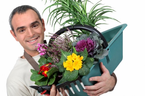 A smiling man holding a black plastic plant pot filled with a variety of colorful flowering plants, including purple, yellow, and red blooms, with green foliage. Behind him, there is a turquoise watering can and a small section of a well-maintained garden, featuring a lush green plant with long, narrow leaves. The scene appears to be outdoors on a clear day, with natural lighting highlighting the vibrant colors of the plants and the neatly arranged garden space, indicative of professional gardening and landscaping work that Lawn Mowing Kensington might provide in the Kensington area. The background hints at a tidy, landscaped garden environment suitable for lawn and garden care services, emphasizing the attention to detail in outdoor maintenance and plant arrangement.